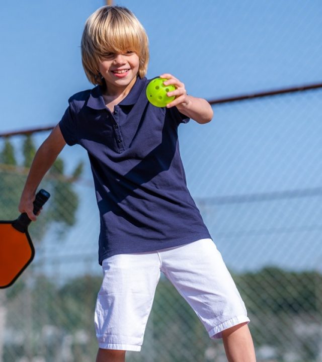 Happy blonde boy playing pickleball game, hitting pickleball yellow ball with paddle, outdoor sport leisure kids activity